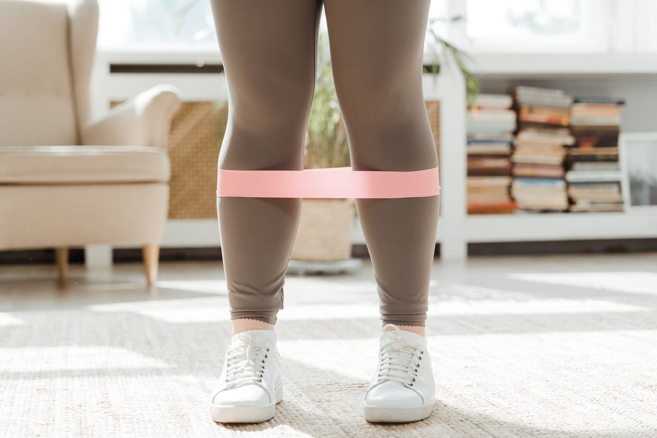 Legs in gray leggings and white sneakers with a pink loop band just above the knees, standing on a rug