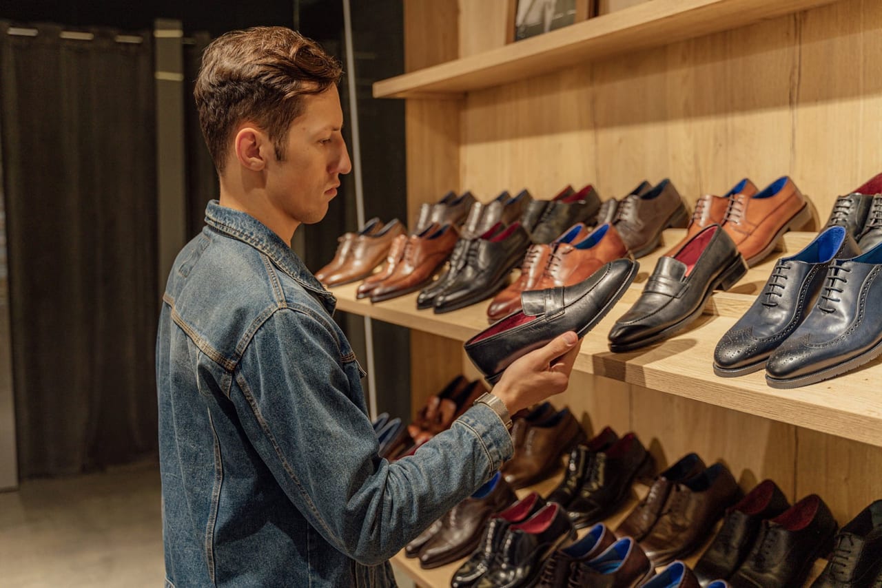 Man in a denim jacket examining a black leather loafer beside a shelf of shoes