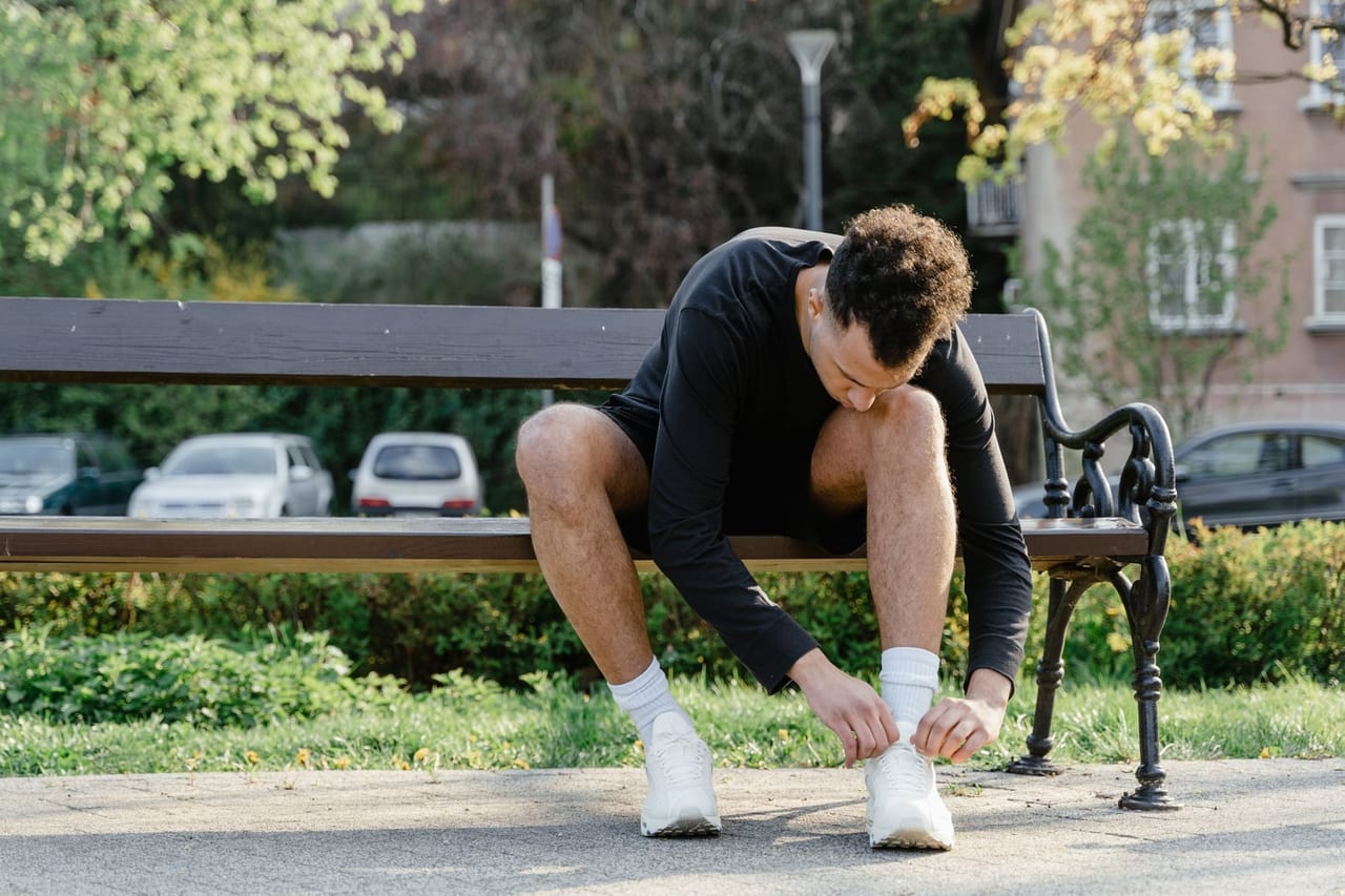 Man sitting on a park bench in sunlight, bending to the laces of his white sneakers