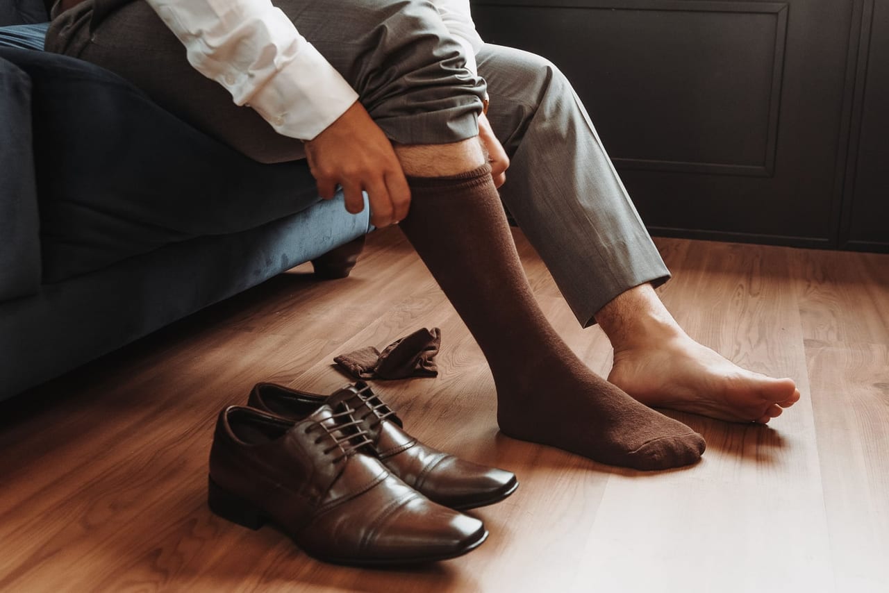 Man on a sofa pulling on a brown sock beside leather shoes on a wooden floor