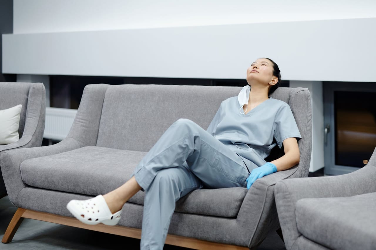 Nurse in light blue scrubs and white clogs leaning back on a gray sofa, eyes closed