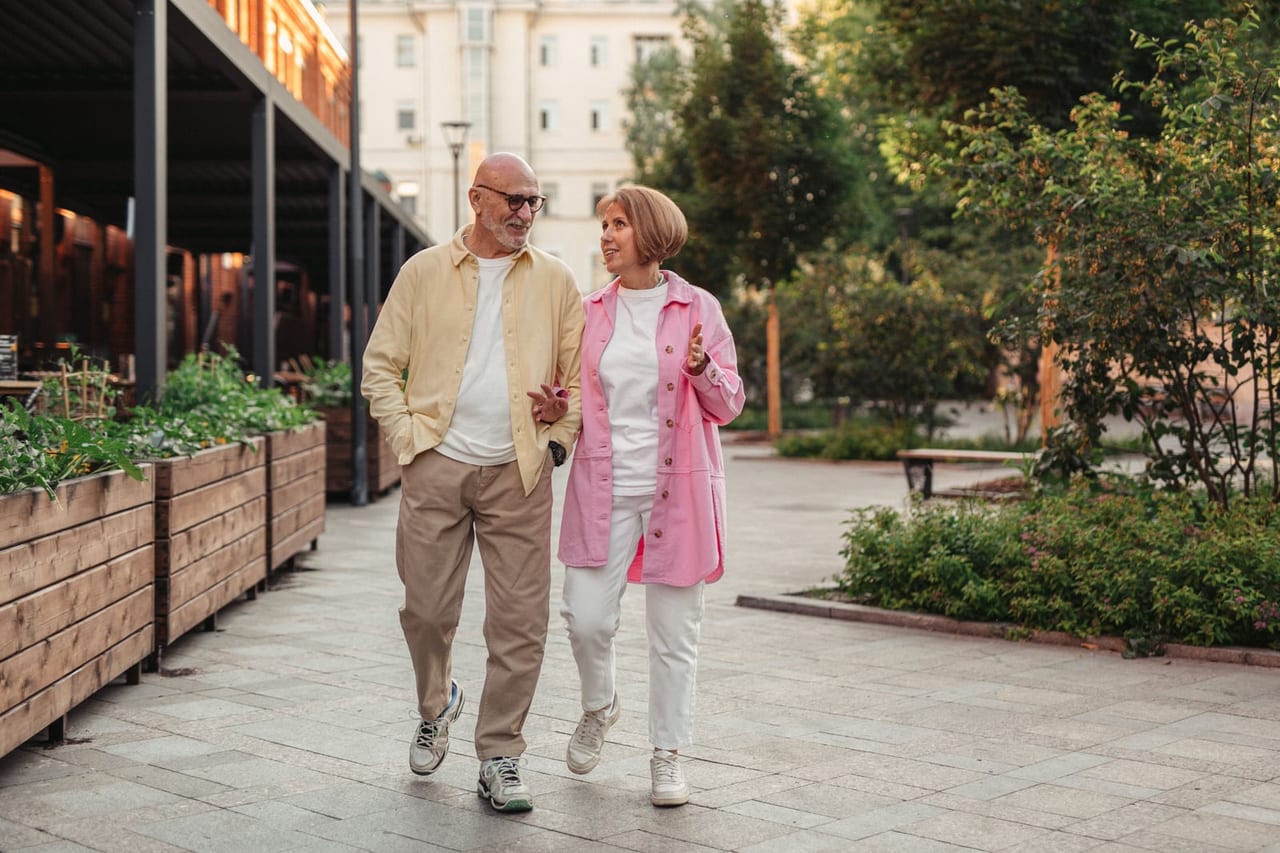 Older couple in sneakers walking and talking along a paved walkway lined with planters