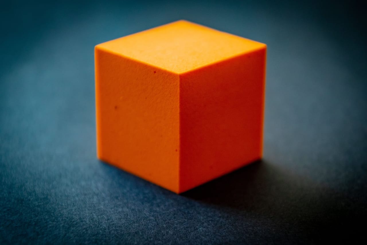 A single orange foam cube on a dark blue surface under soft light