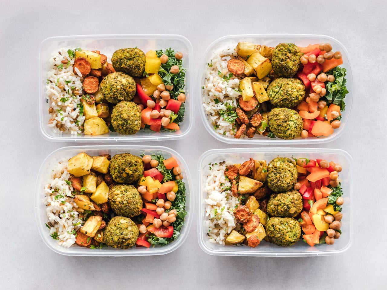 Four clear containers of rice, falafel, chickpeas, roasted vegetables and greens on a gray surface