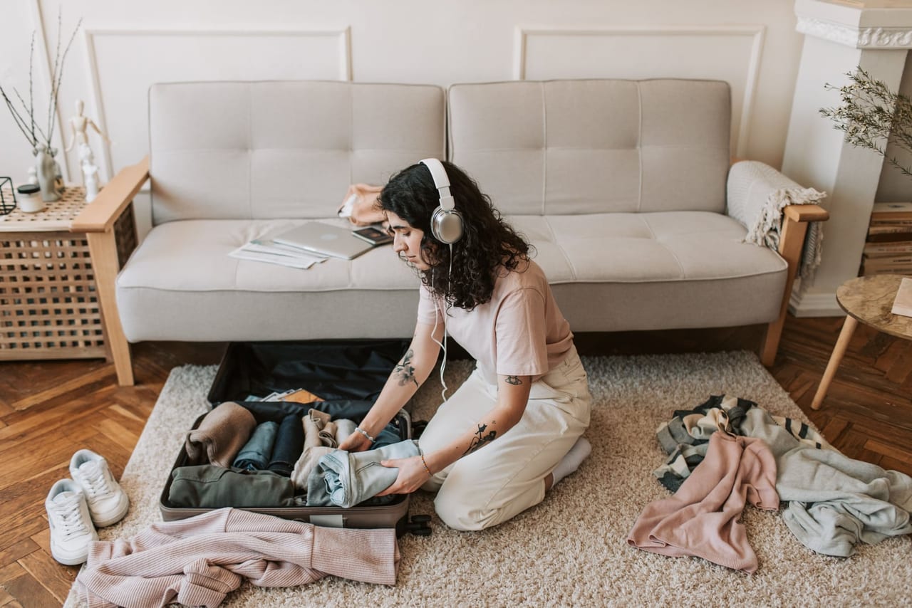 Woman packing rolled clothes into a suitcase on a rug, with white sneakers set beside it