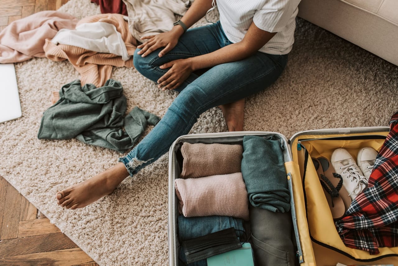 Woman sitting barefoot on a rug beside an open suitcase packed with clothes and white sneakers