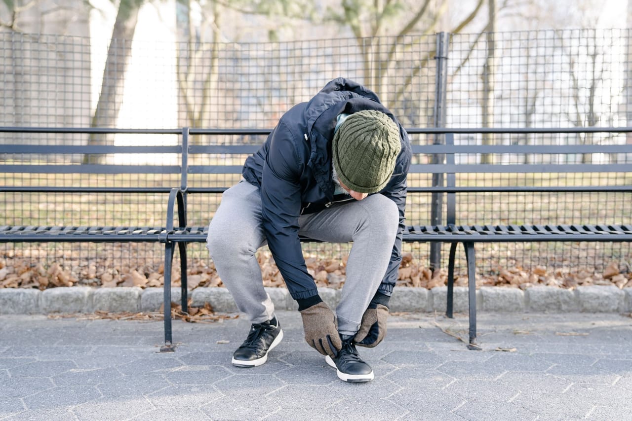 Man in a knit hat and gloves on a park bench, tying a sneaker lace