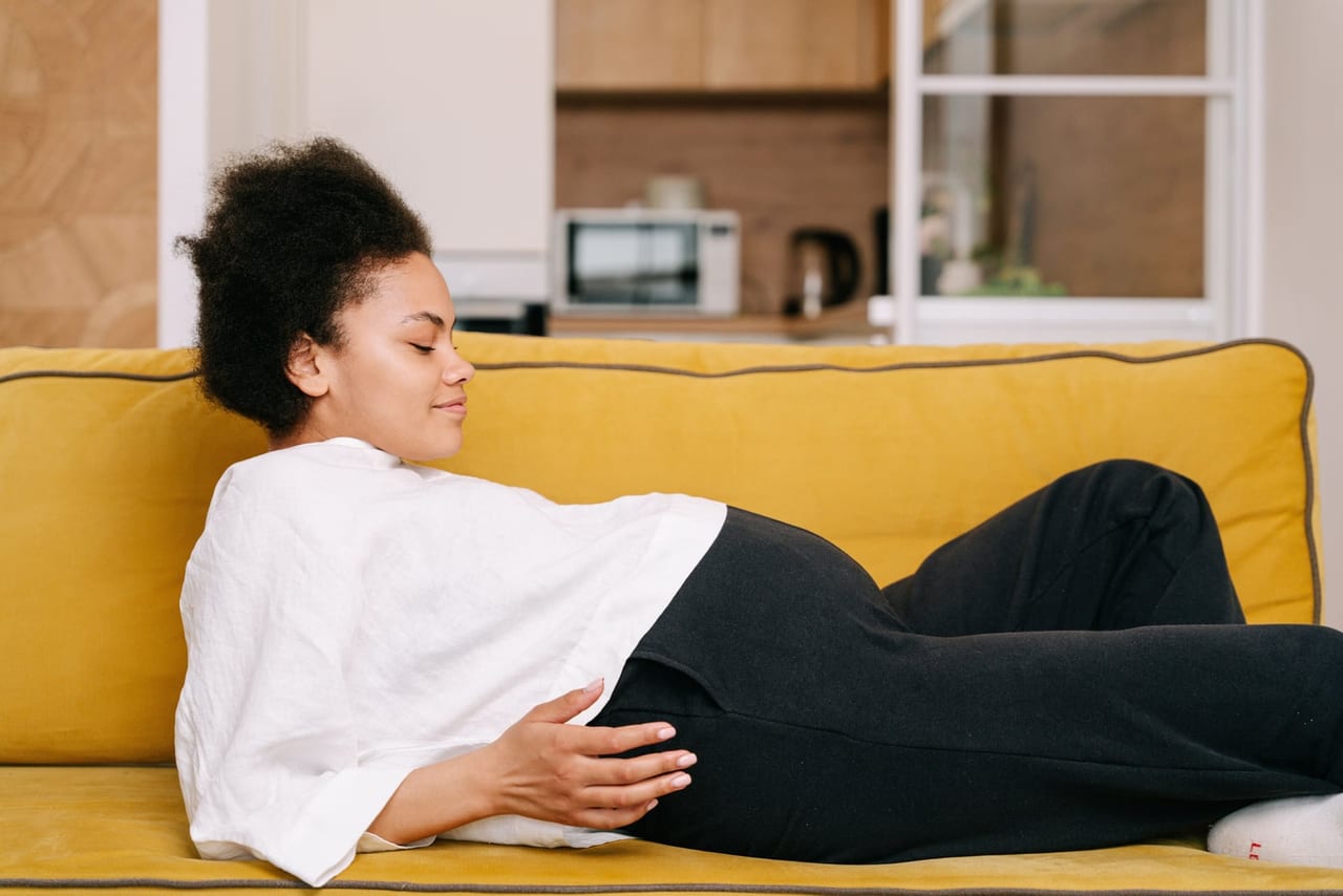 Pregnant woman resting on a yellow sofa with her legs up, one hand on her belly
