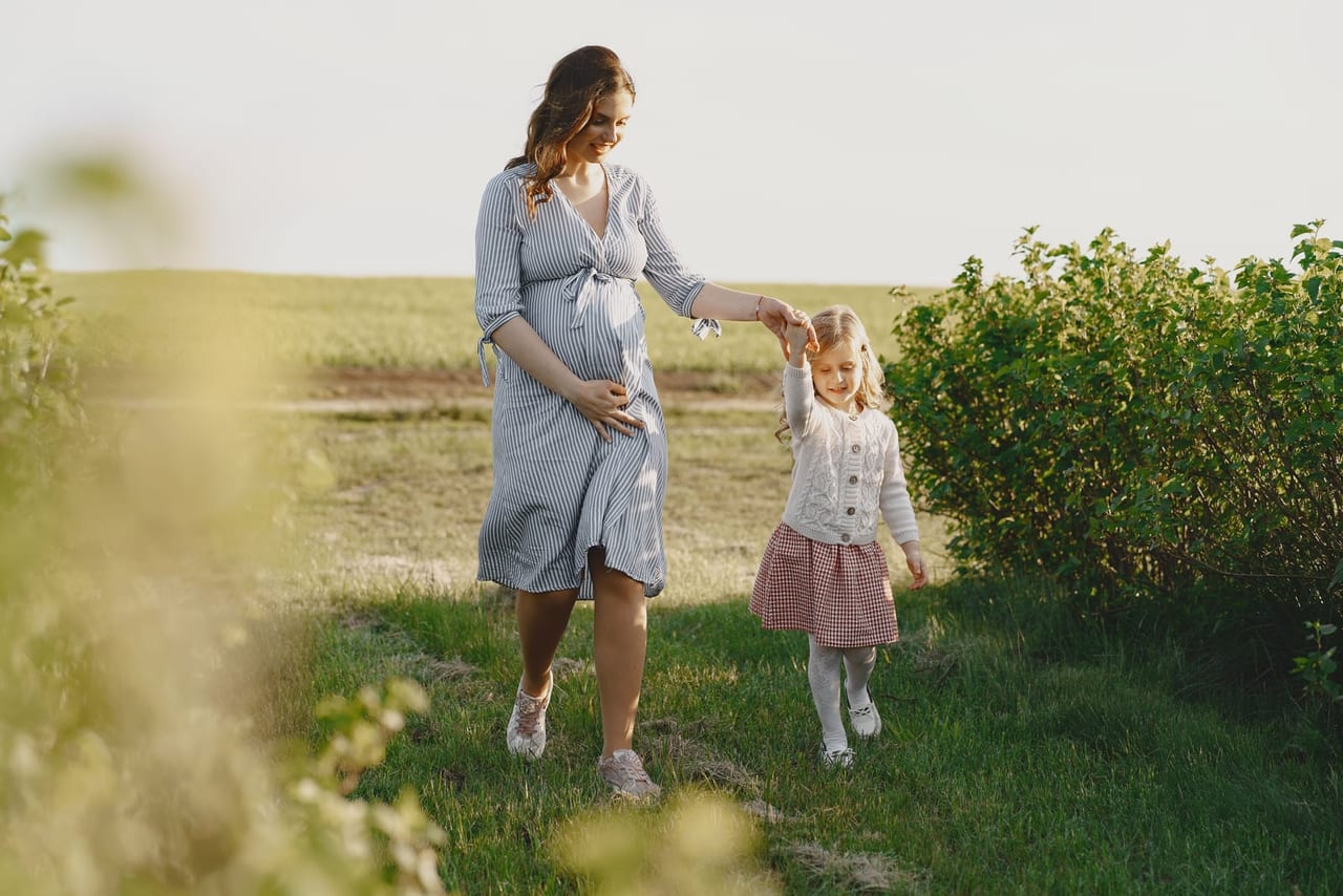 Pregnant woman in a striped dress and sneakers walking across grass, holding a girl's hand