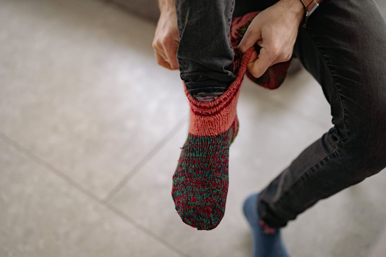 Man in dark jeans pulling a red and green knit sock onto his foot
