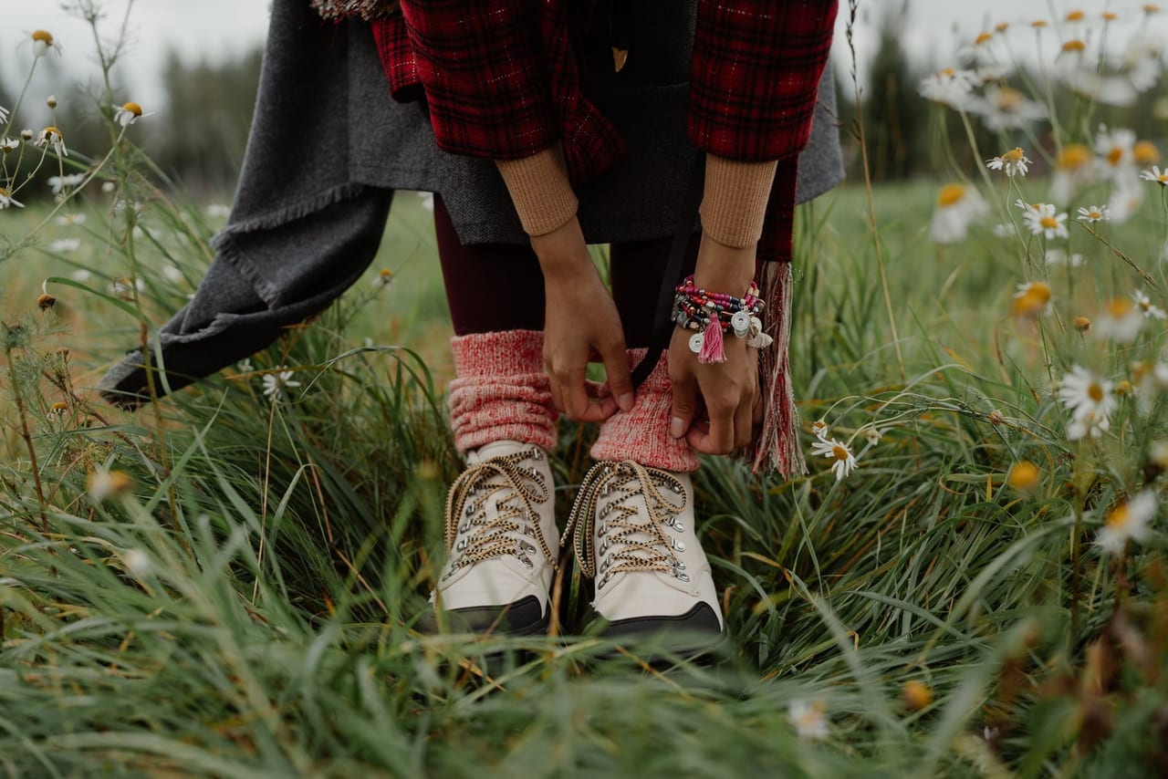 Hiker in a plaid jacket pulling up wool socks over her boots in a meadow