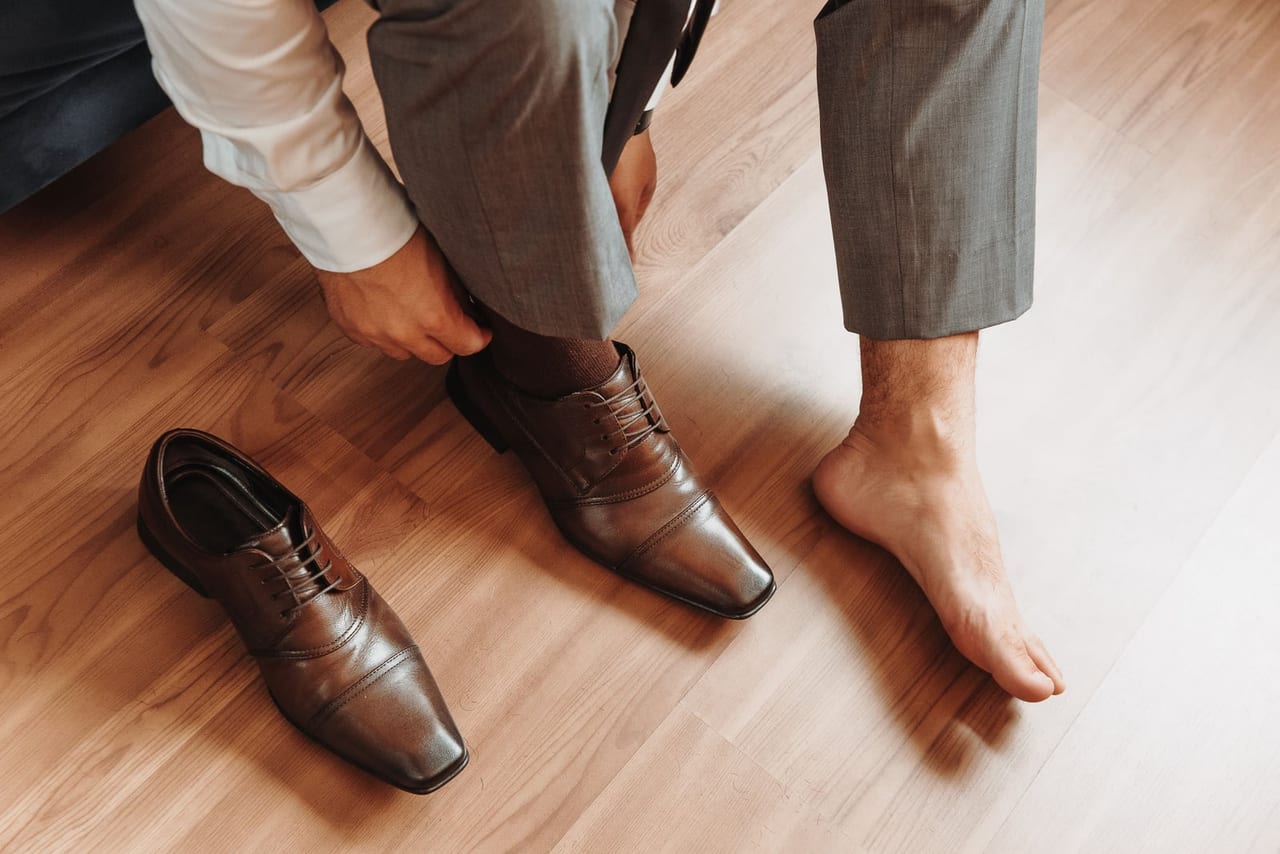 Man in a gray suit putting on brown leather shoes, one foot still bare