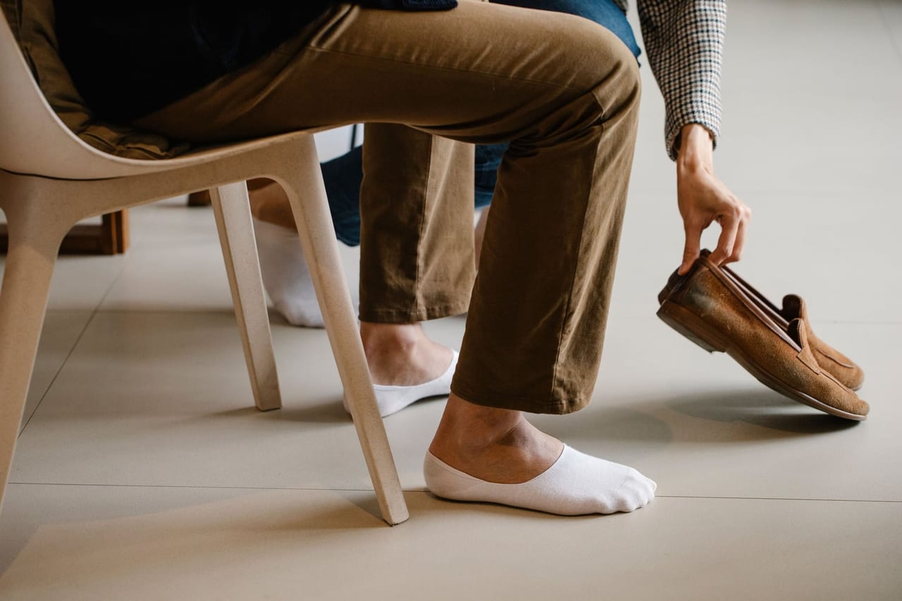 Seated man in no-show socks reaching for a pair of brown suede loafers