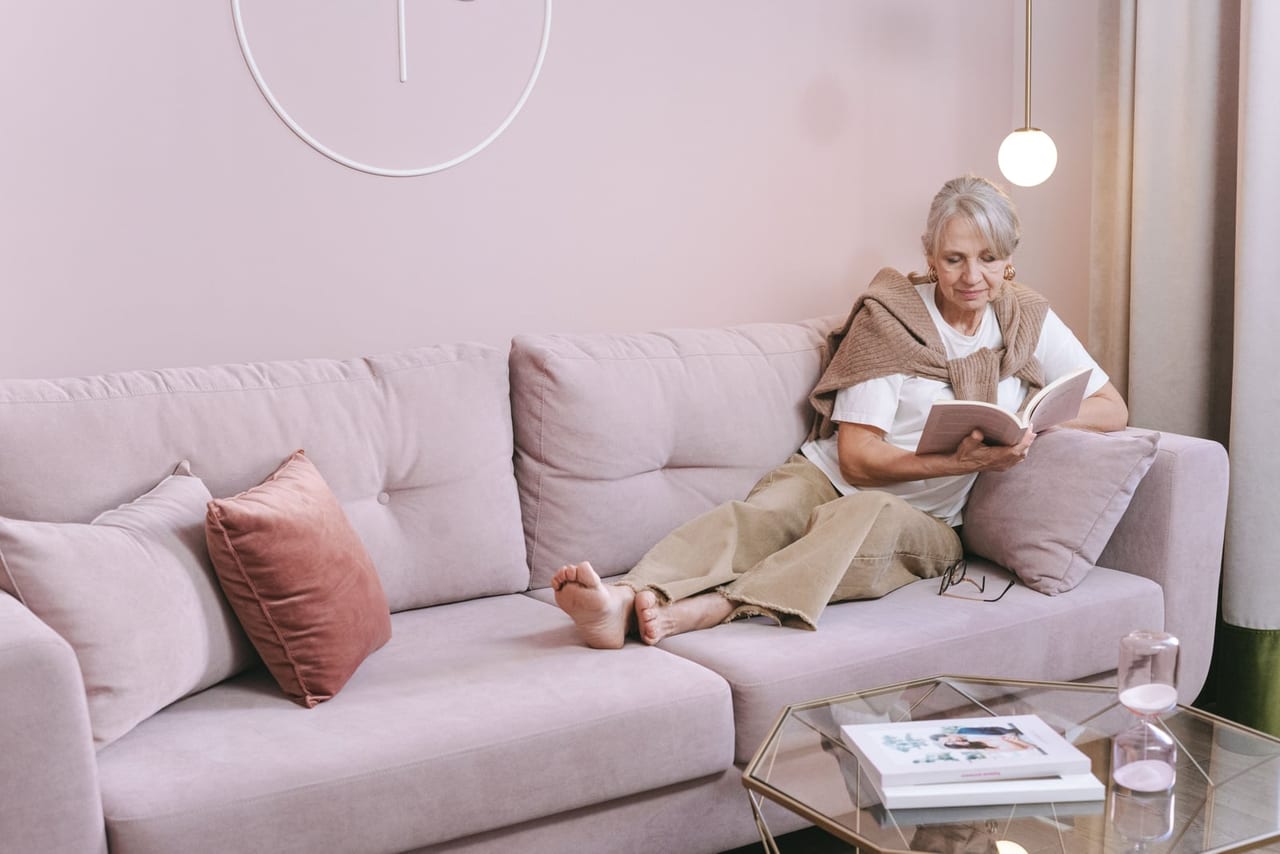 Older woman reading a book on a pale pink sofa with her bare feet up