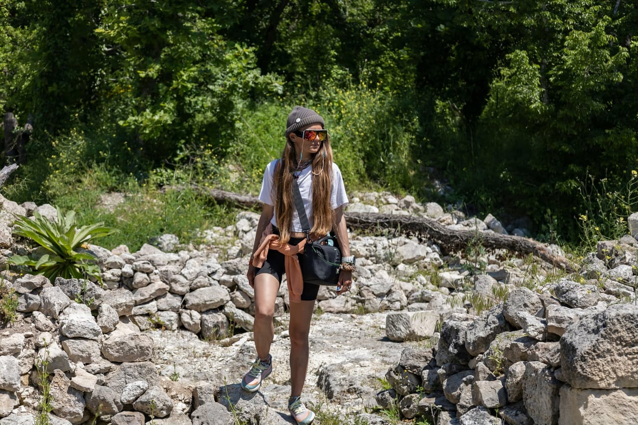 Woman in hiking shoes and a beanie walking along a rocky path through green woods