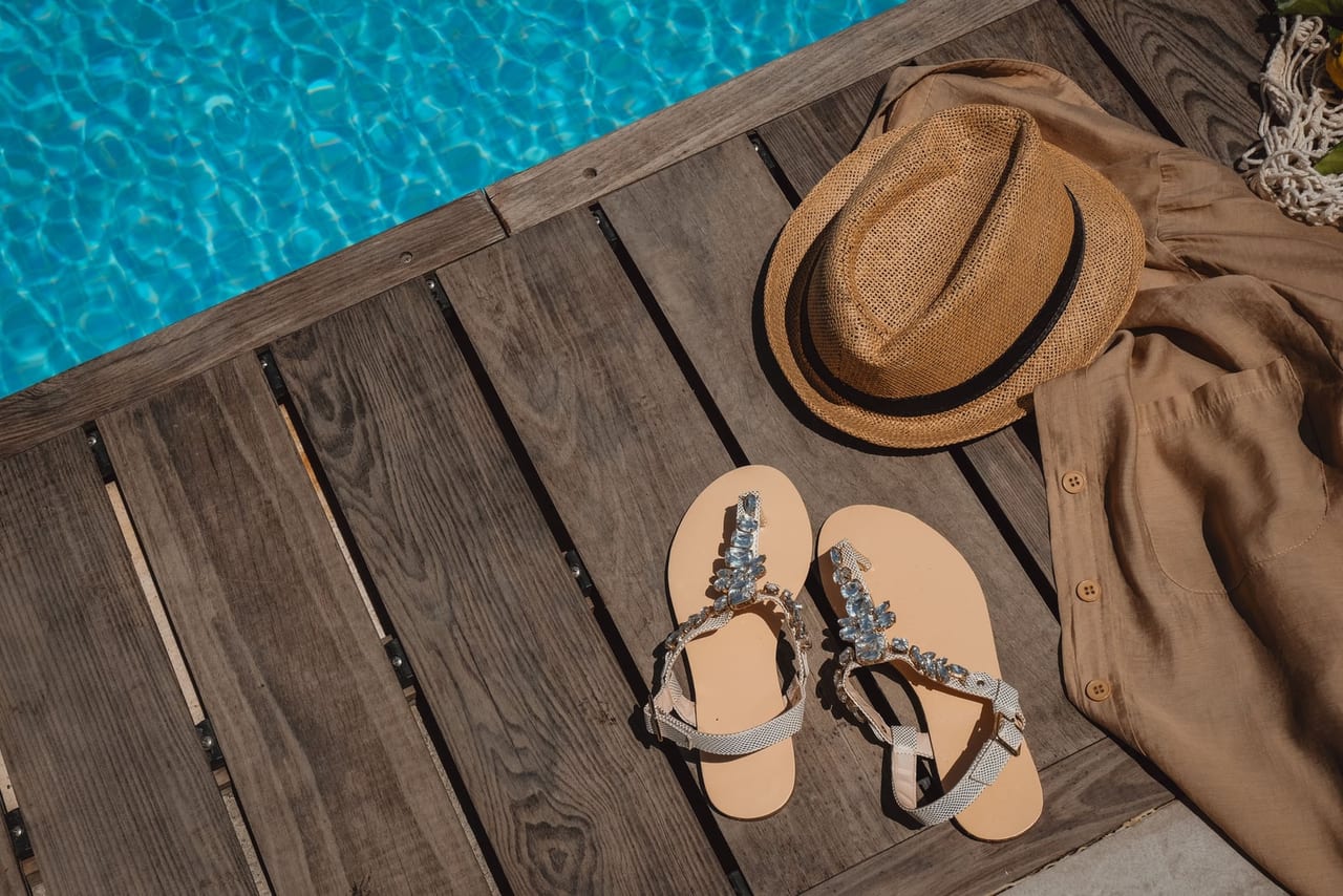 Embellished sandals and a straw hat on a wooden deck beside a bright blue pool