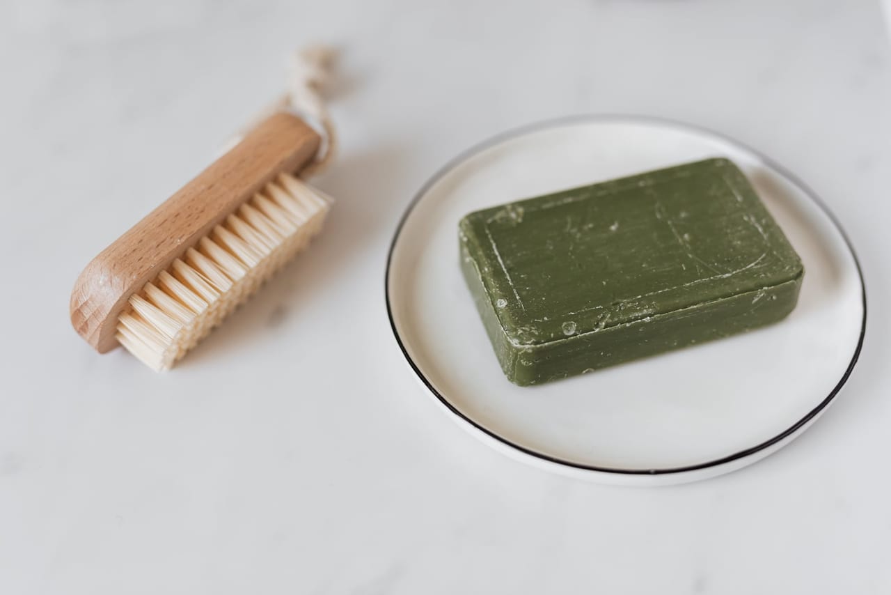 Wooden scrub brush beside a green bar of soap on a white plate on marble