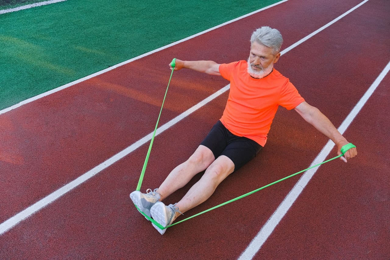 Older man seated on a running track pulling a green band looped around his feet