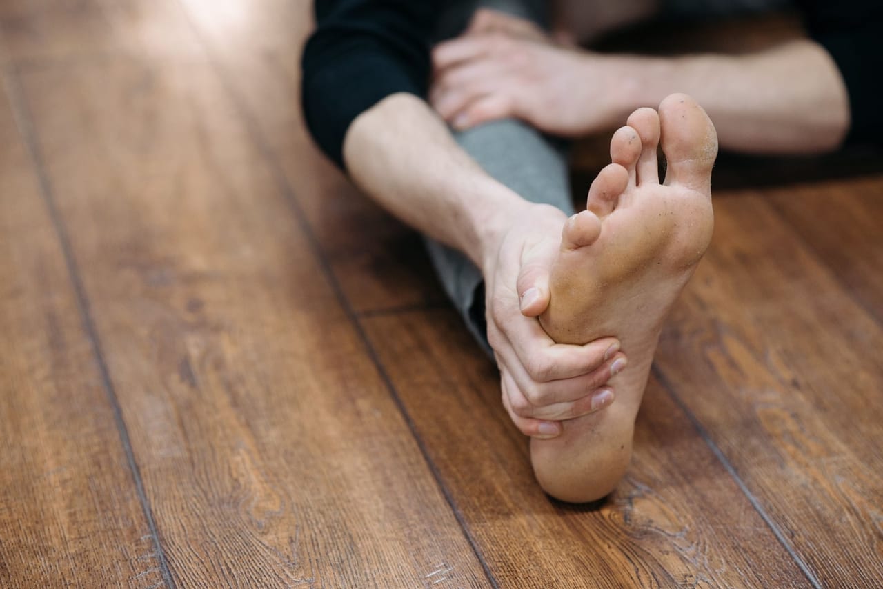 Person seated on a wooden floor holding a bare foot in a seated stretch