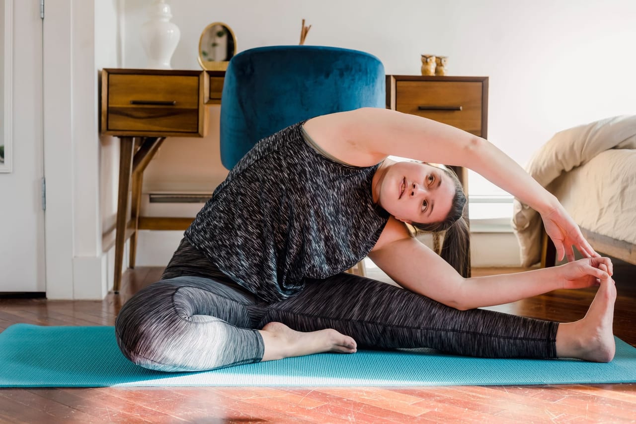 Woman on a teal mat in a bedroom doing a seated side stretch toward one foot