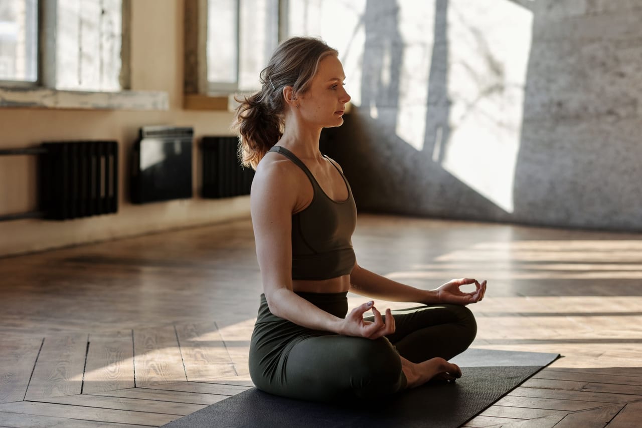Woman sitting cross-legged on a black mat in a sunlit room, hands resting on her knees
