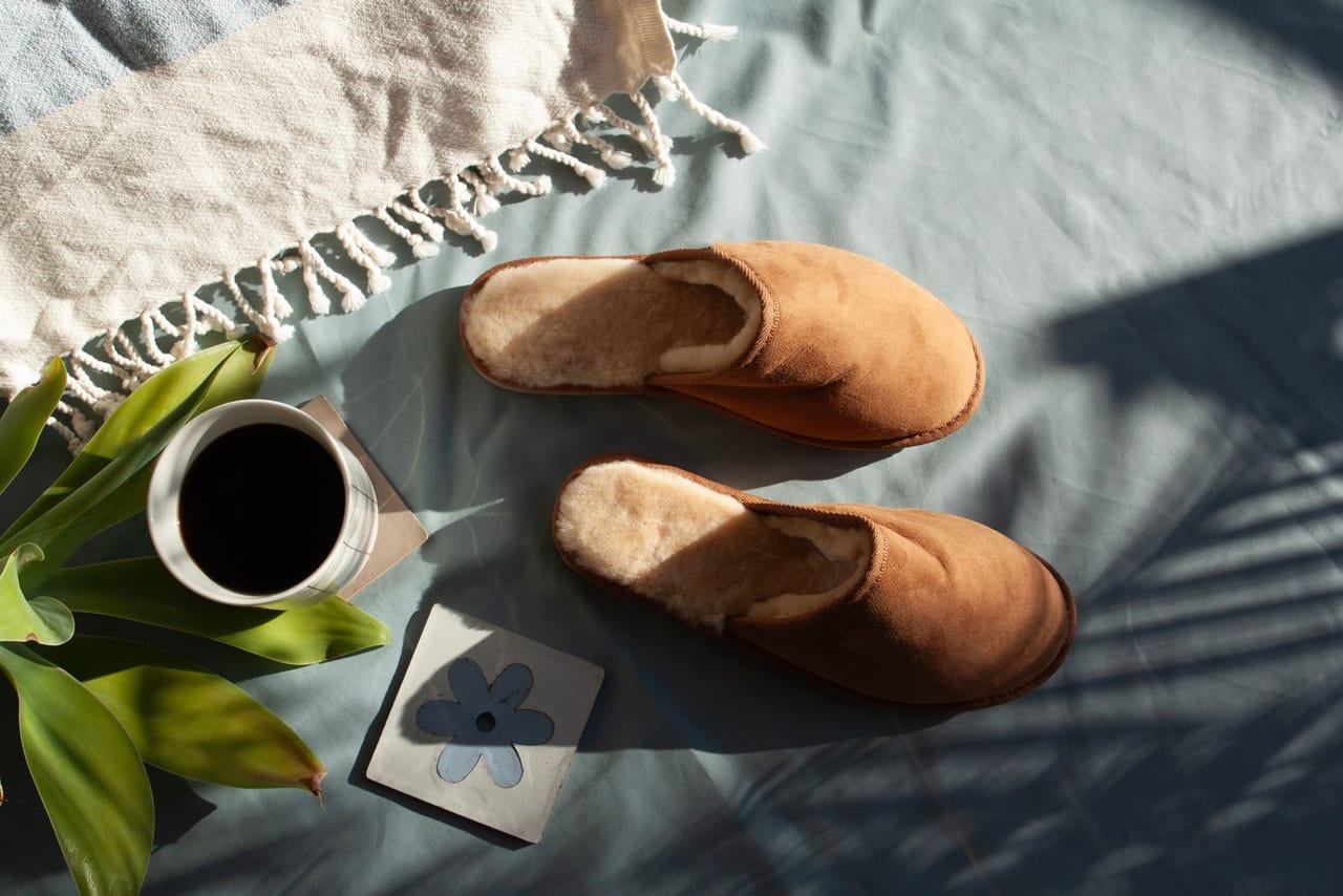 Sheepskin-lined suede slippers on a blue bedsheet in sunlight beside a coffee cup and a plant