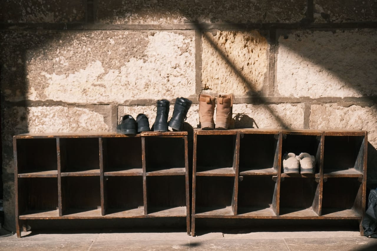 Boots and shoes on weathered wooden cubby racks against a sunlit stone wall