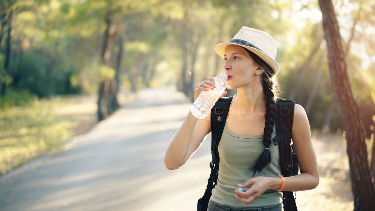 Woman in a straw hat and backpack sipping water on a sunny, tree-lined path