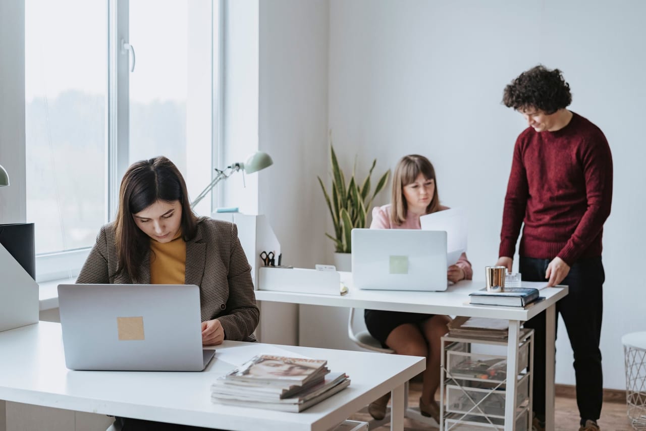 Two women working at laptops while a colleague stands at a desk in a bright office