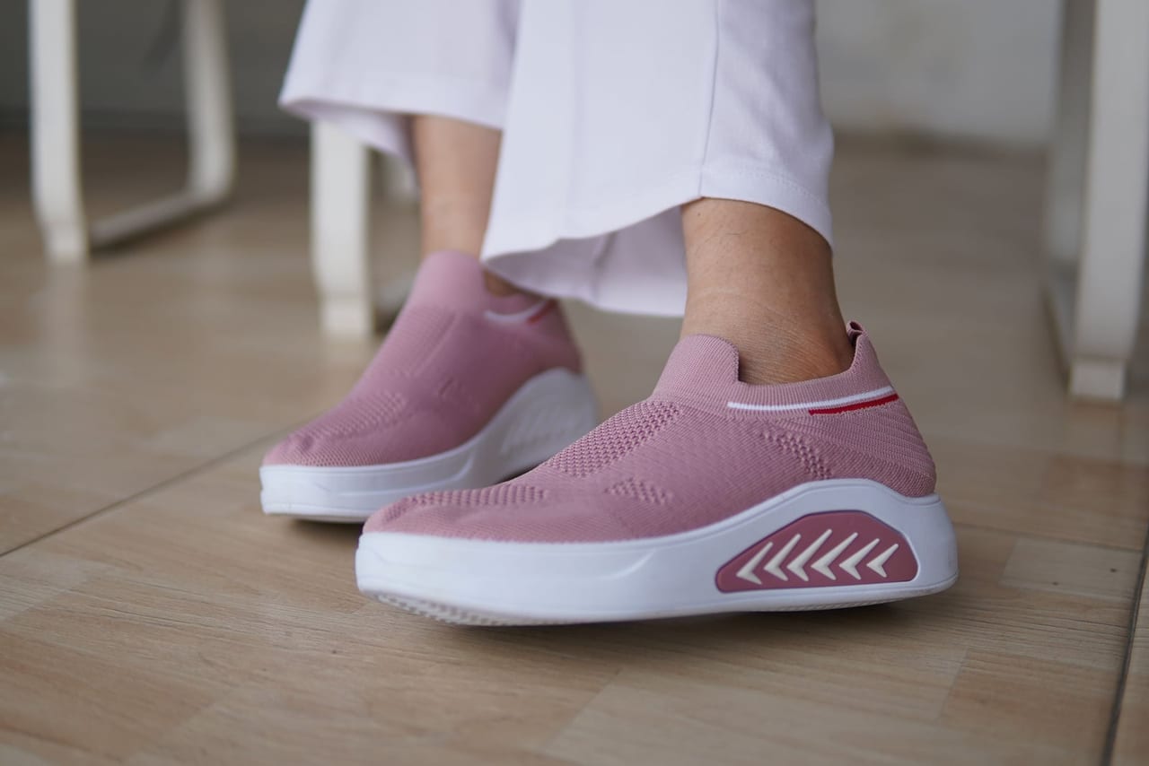 Pink knit slip-on sneakers with thick white soles worn indoors on a wood-look floor