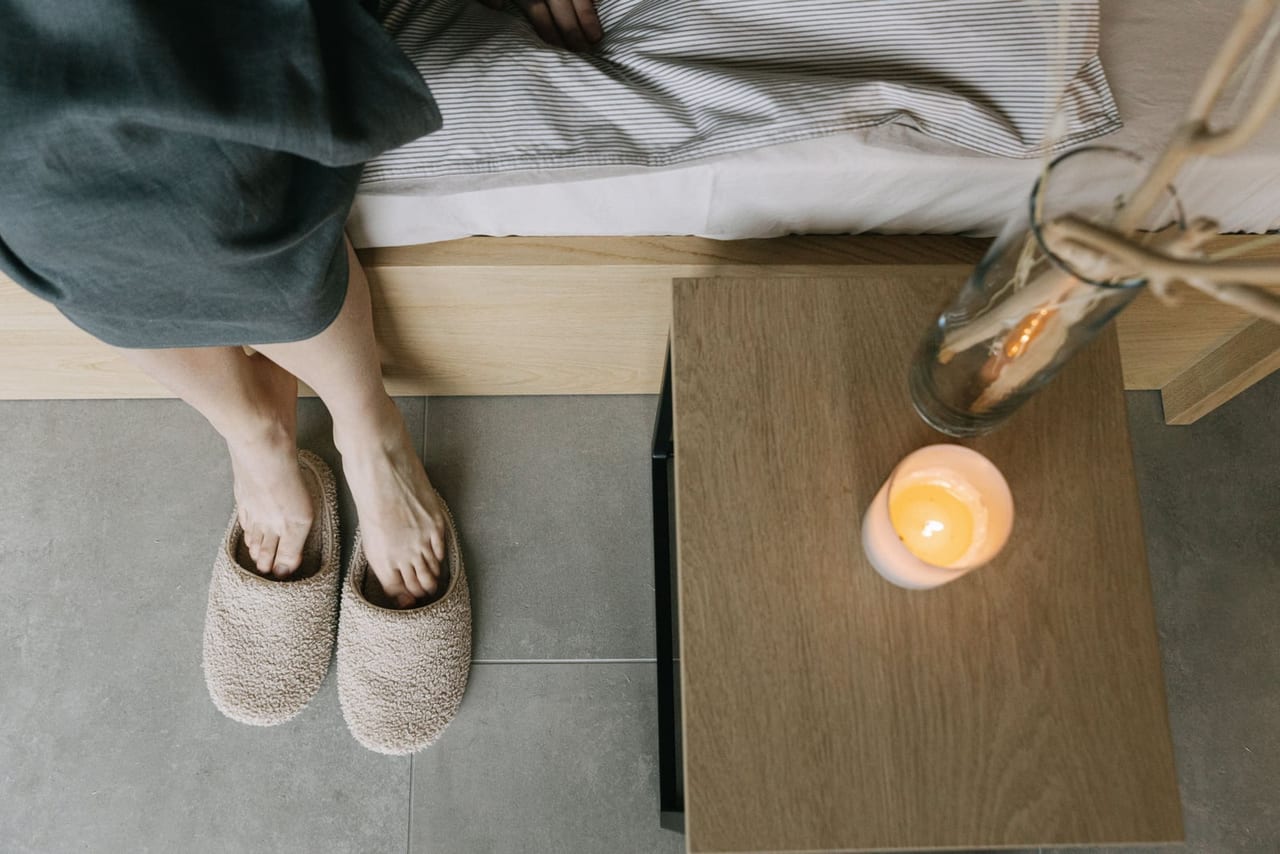 Bare feet slipping into fleecy beige slippers beside a bed and a lit candle, from above