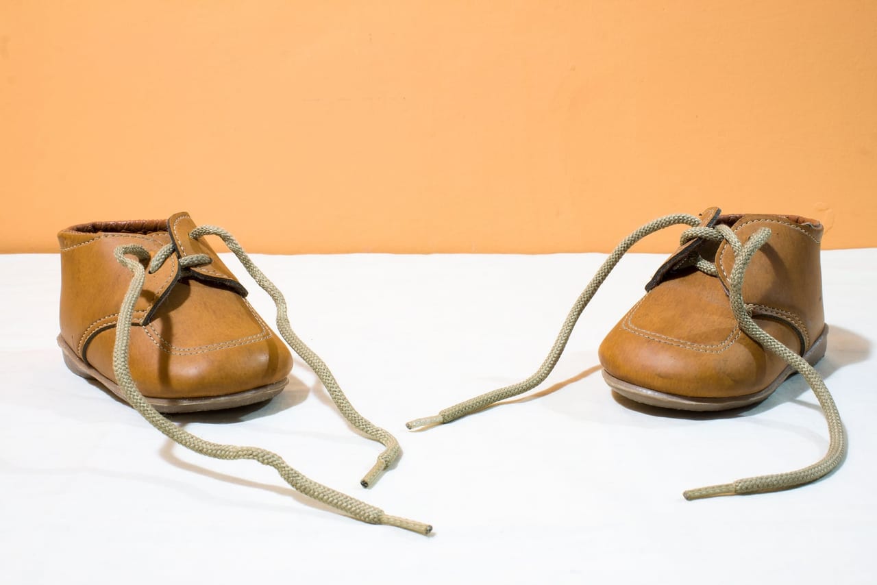 A pair of small brown leather children's shoes with loose laces against an orange wall