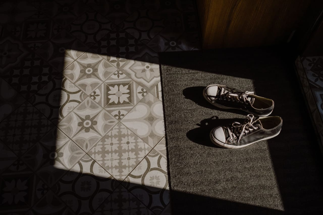 Pair of dark sneakers on a doormat in a patch of sunlight next to tiles
