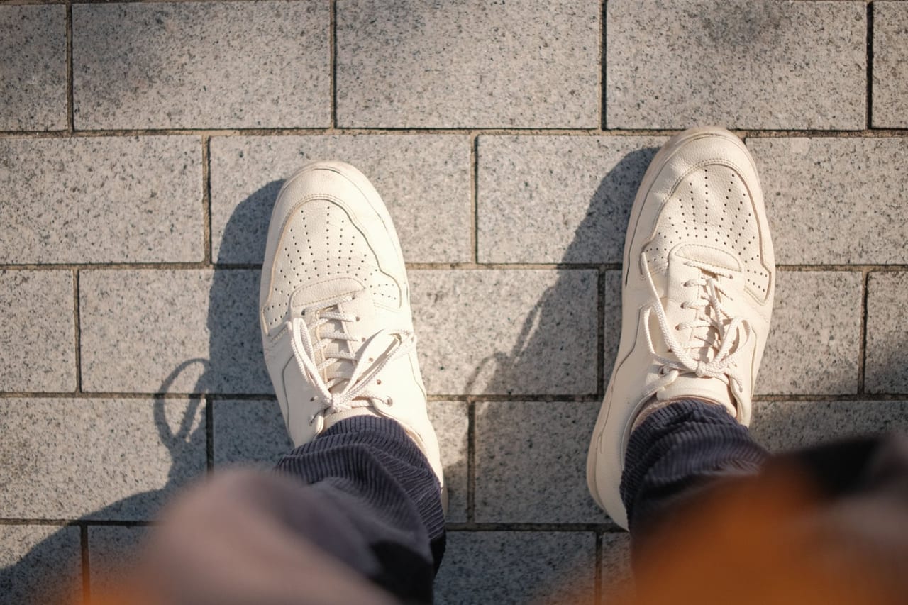 Looking down at off-white leather sneakers on gray stone paving in low sun
