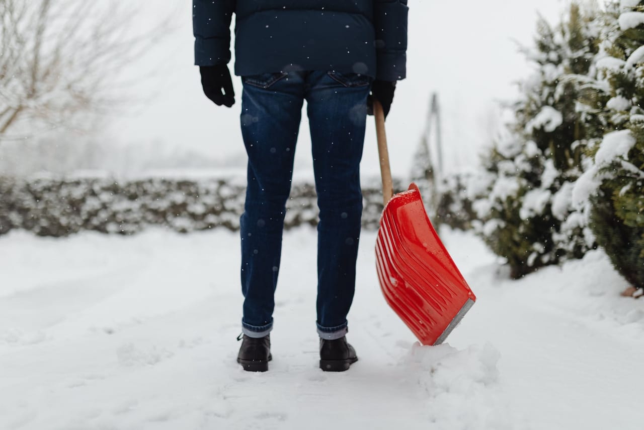 Person in jeans and winter boots pausing on a snowy path with a red snow shovel