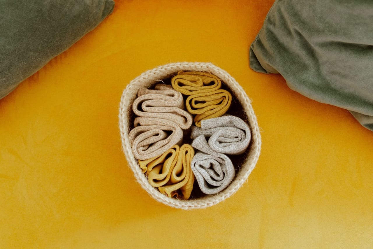 Rolled socks in beige, mustard and gray in a small knit basket on a yellow surface