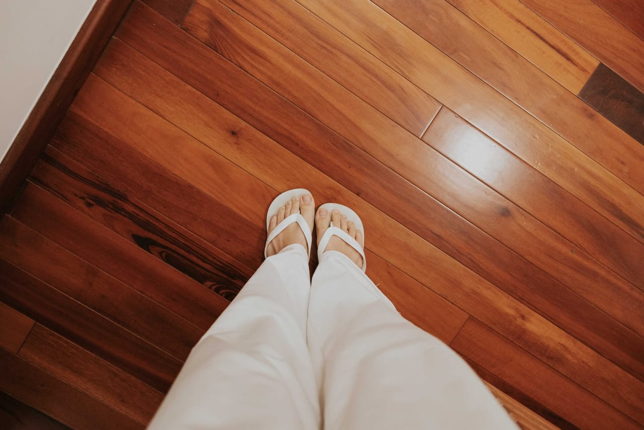 Looking down at white sandals and cream trousers on a polished wooden floor