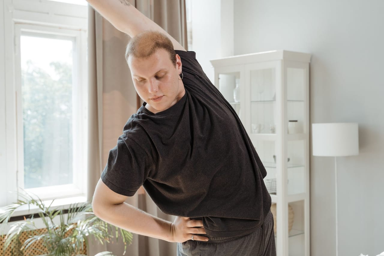 Man in a dark T-shirt doing a side stretch beside a window in a bright room