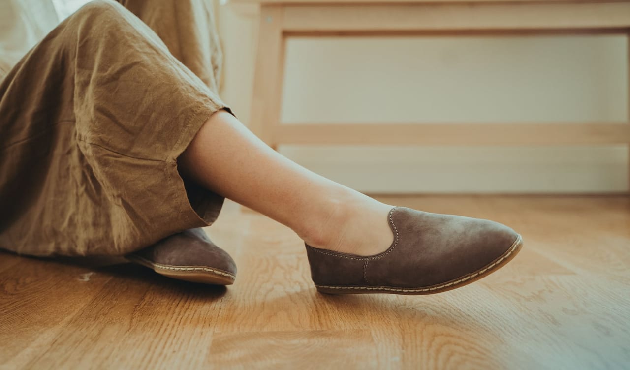 Soft brown suede closed-back flats on relaxed feet resting on a sunlit wooden floor