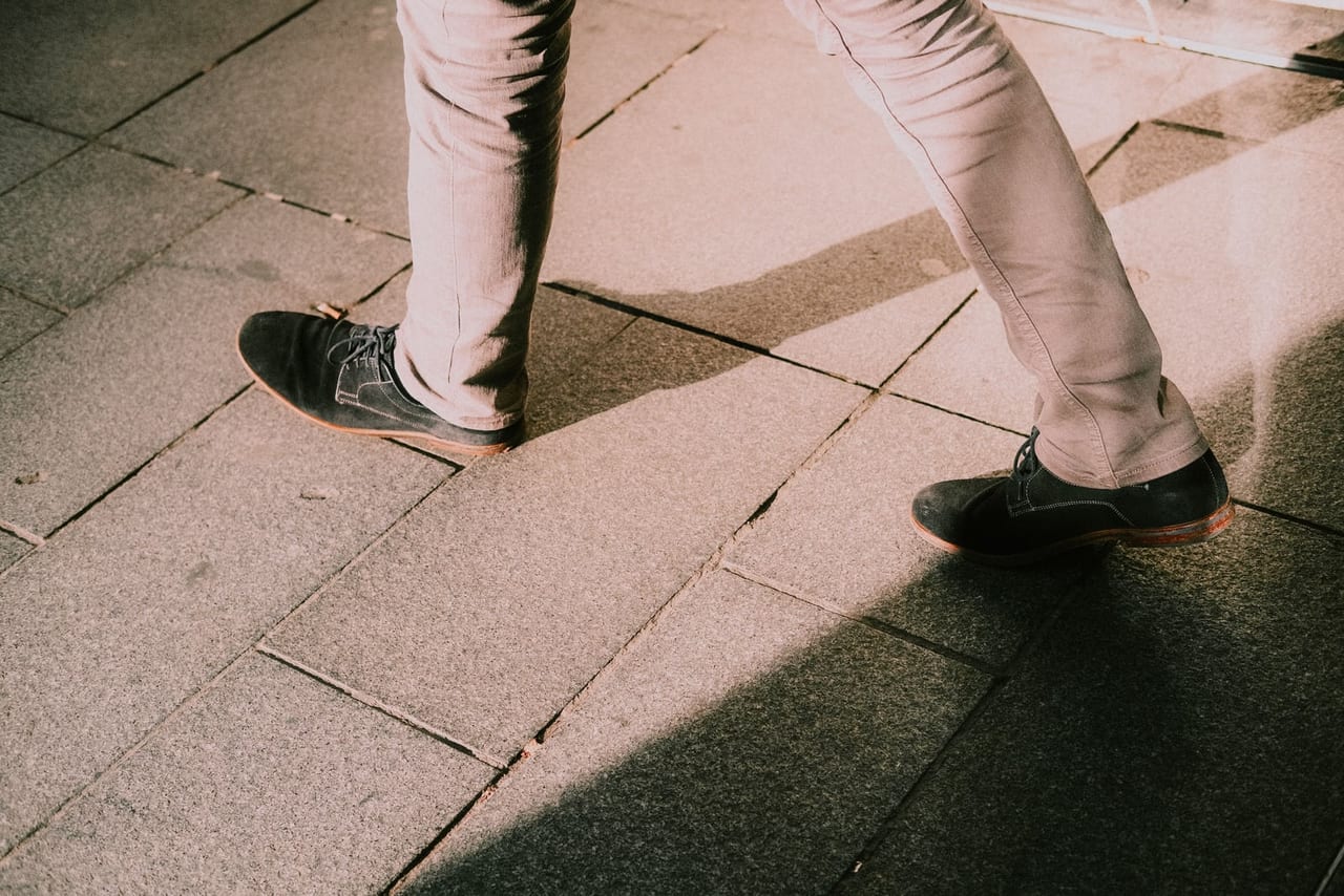 Legs in light trousers and dark suede lace-up shoes walking across sunlit stone paving