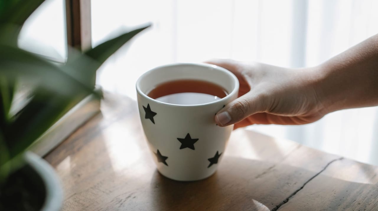 A hand resting on a cup of tea on a wooden table beside a window