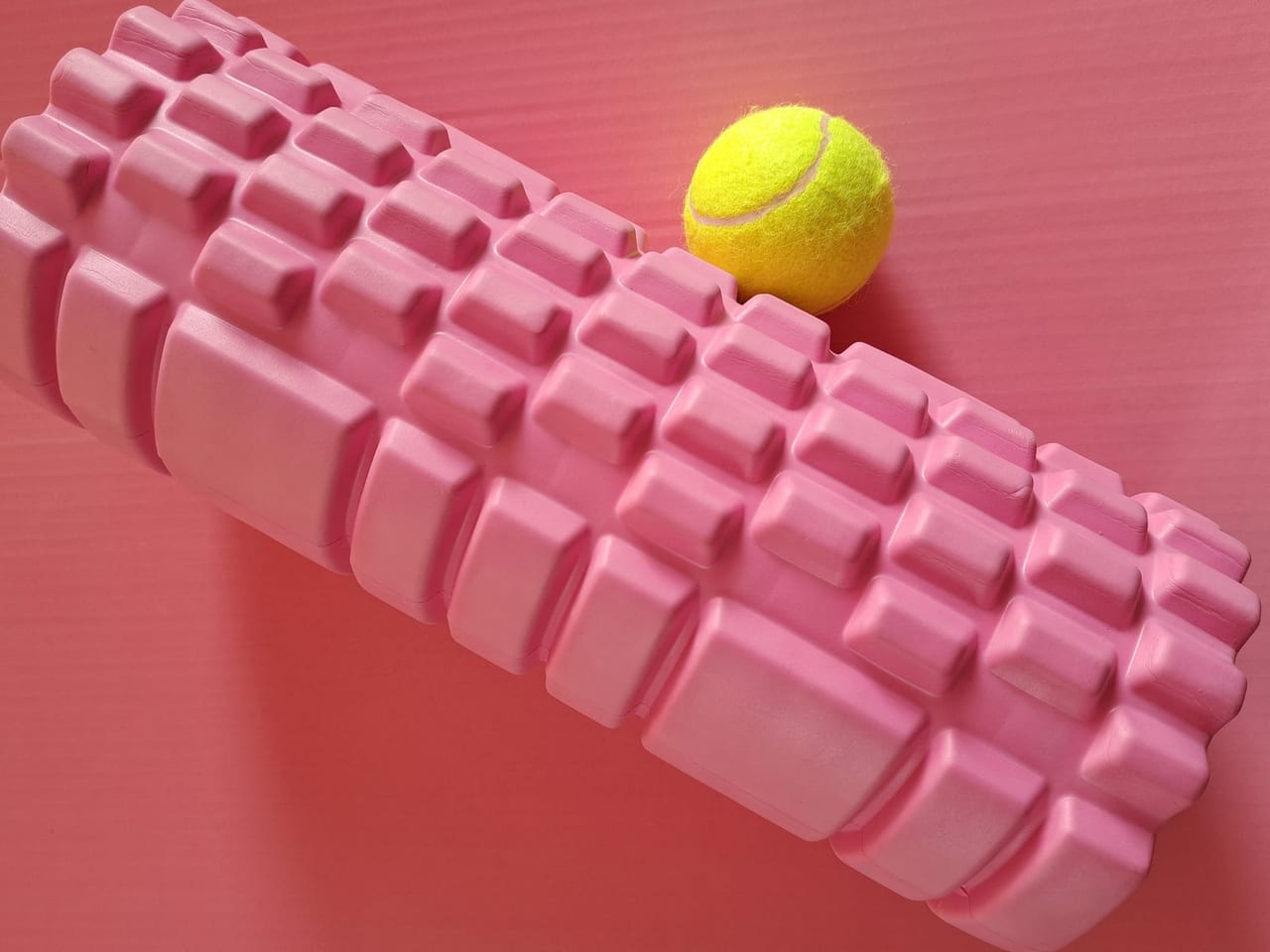 Yellow tennis ball resting beside a pink textured foam roller on a pink mat