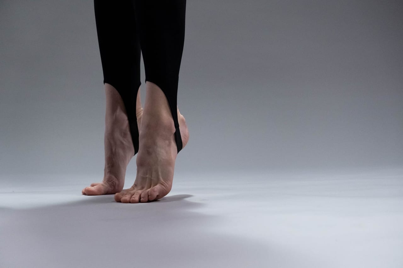 Bare feet in black stirrup leggings rising onto tiptoe on a pale gray studio floor