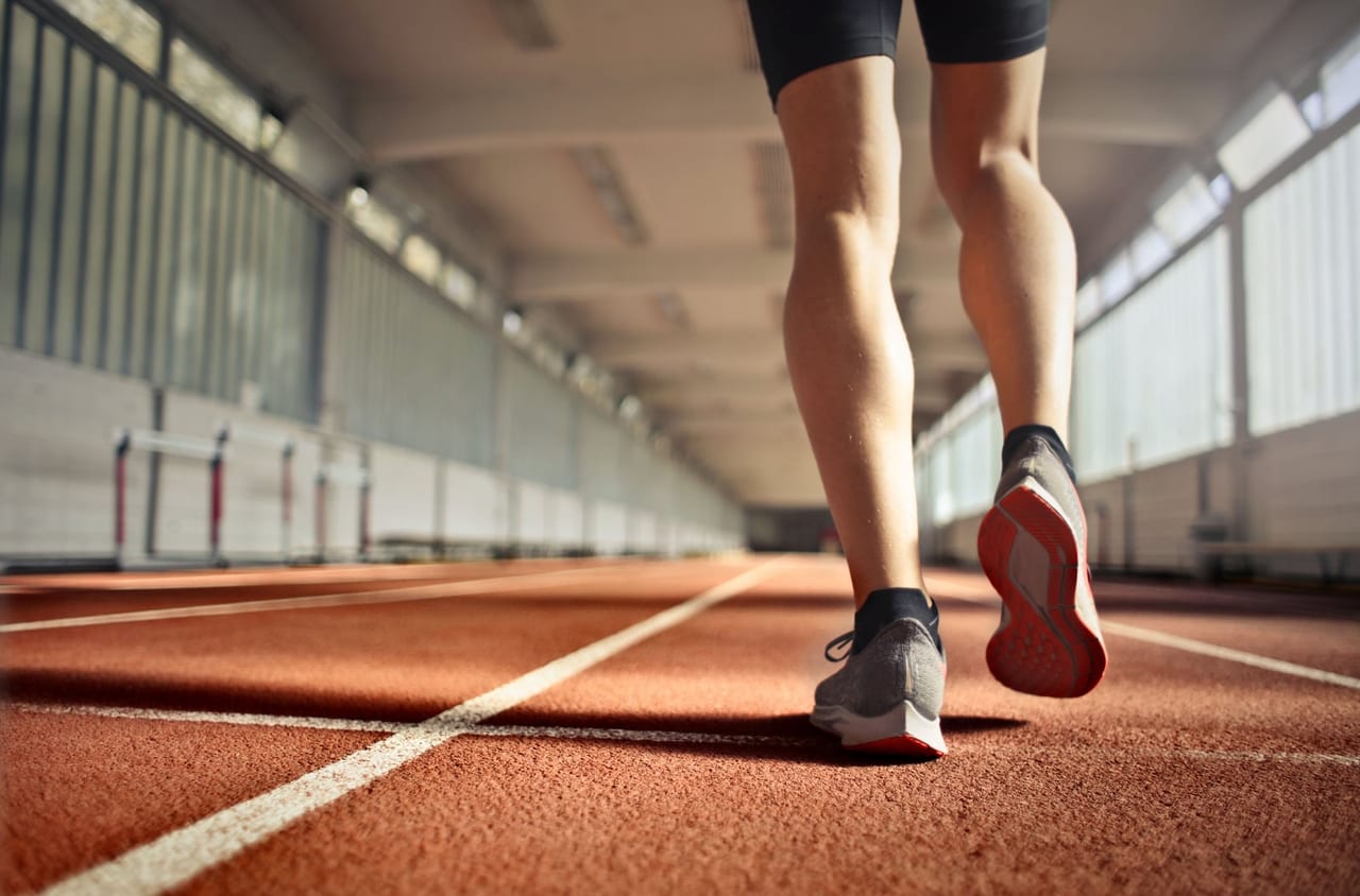 Person in gray running shoes walking along a red indoor track, one heel lifting
