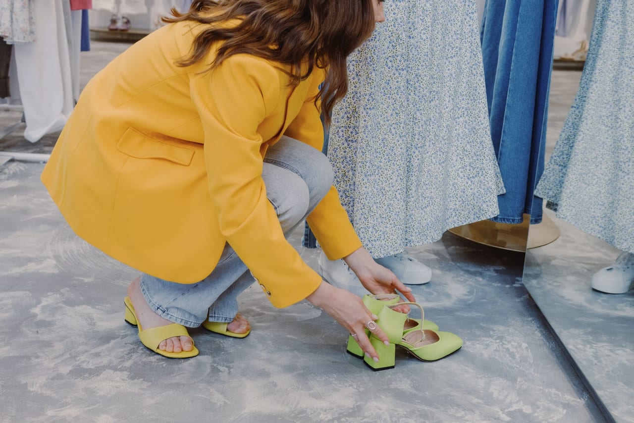 Woman in a yellow blazer crouching in a clothing store to try on green heeled sandals