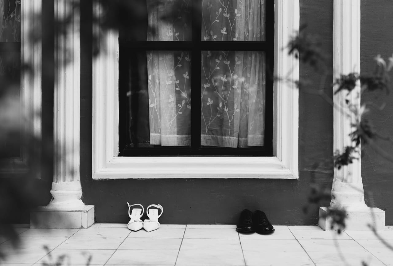 Two pairs of shoes set well apart on a tiled porch below a curtained window