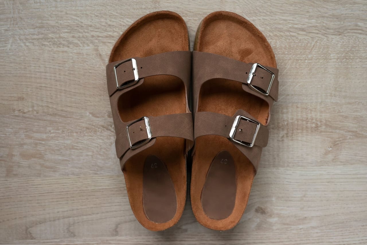 Brown two-strap buckle sandals with cork-edged footbeds, seen from above on a pale wooden floor