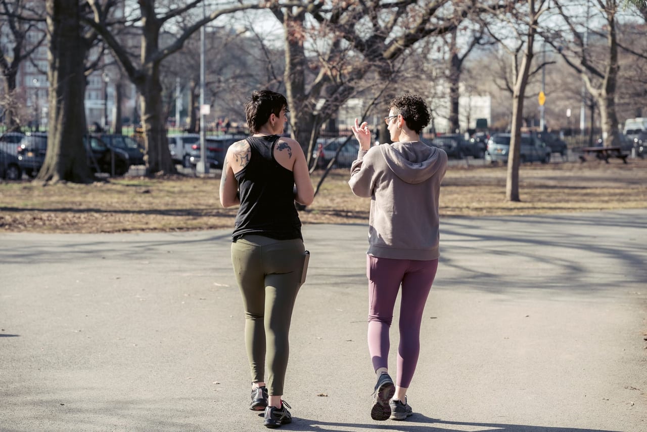 Two women in activewear walking side by side and talking on a sunlit park path