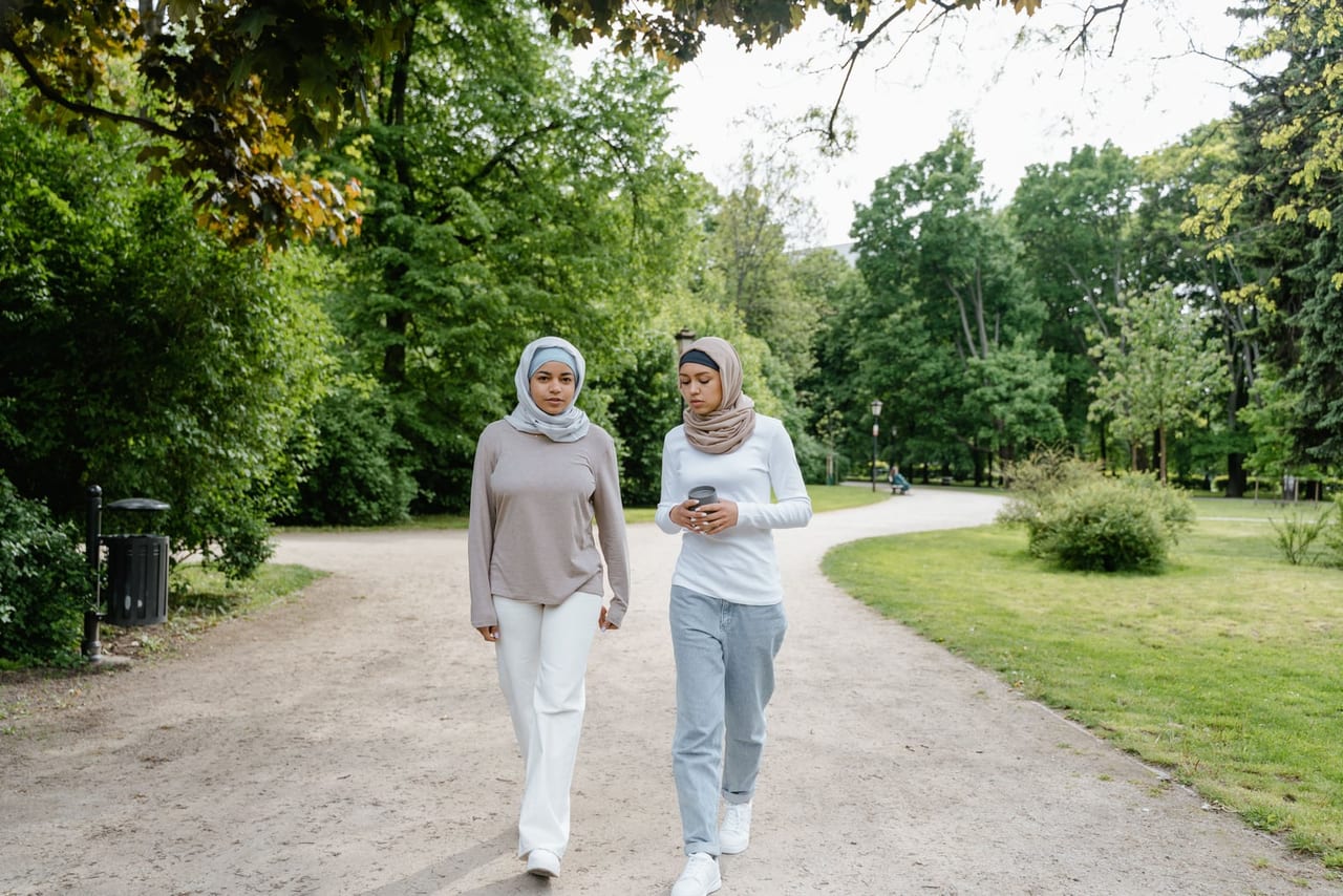 Two women in headscarves walking side by side along a gravel park path lined with trees