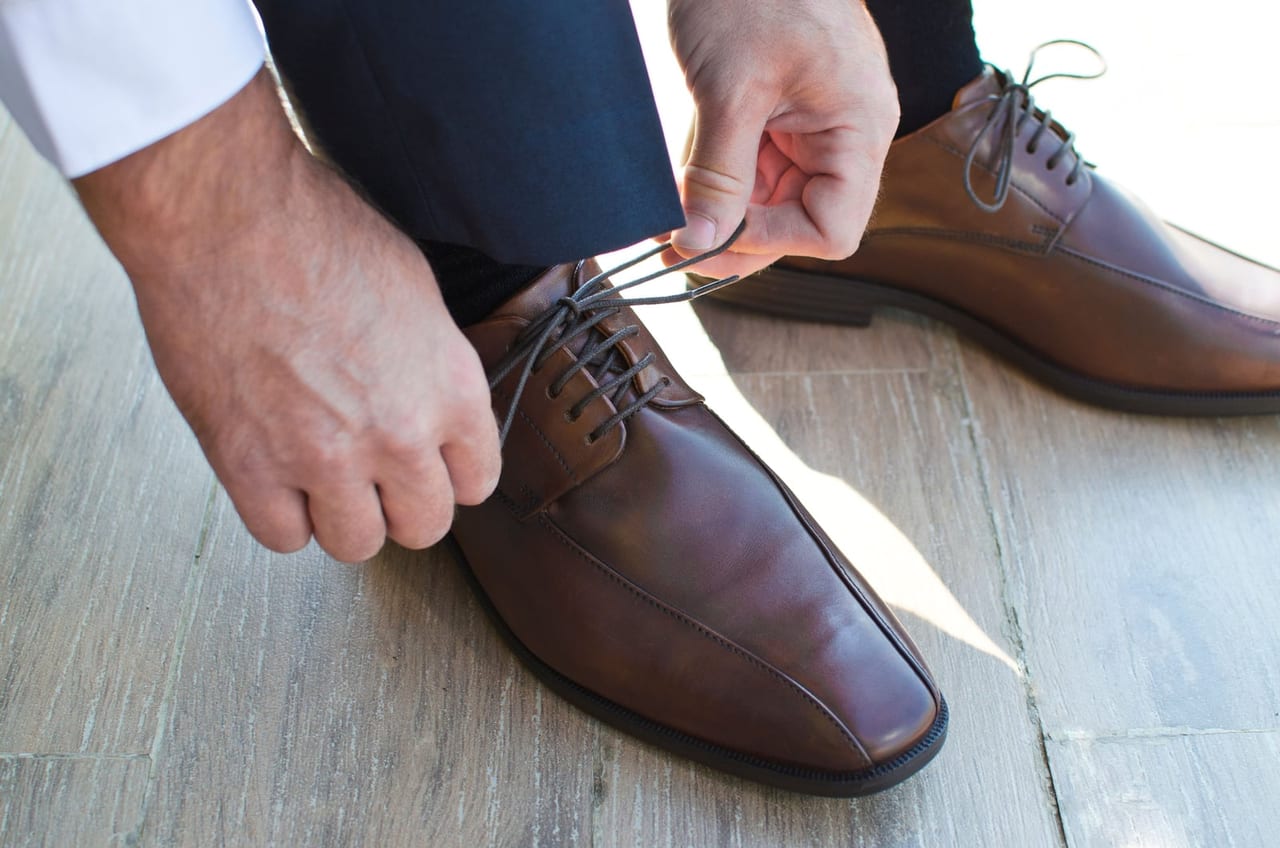 Hands tying the laces of a brown leather dress shoe on a pale wooden floor